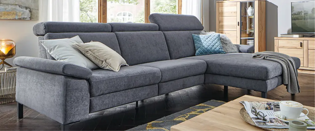 Modernes Wohnzimmer mit grauem Ecksofa, Couchtisch aus Holz und passenden Wohnmöbeln vor großen Fenstern als Beispiel für komfortable Wohnraumgestaltung